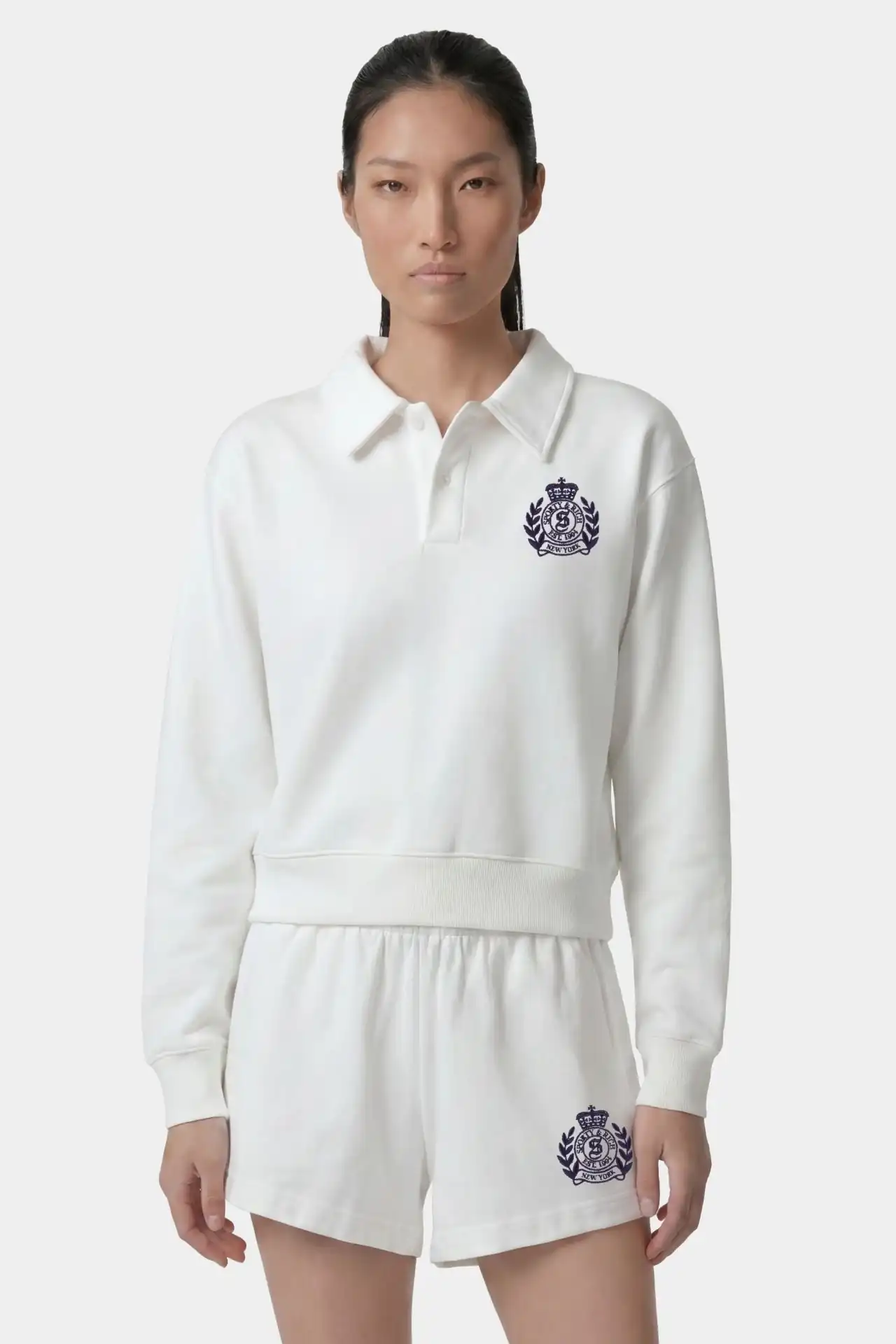 A medium shot shows a woman wearing a white long-sleeved polo shirt and white shorts, both featuring a navy blue crest logo with the text 'SPORTY & RI...