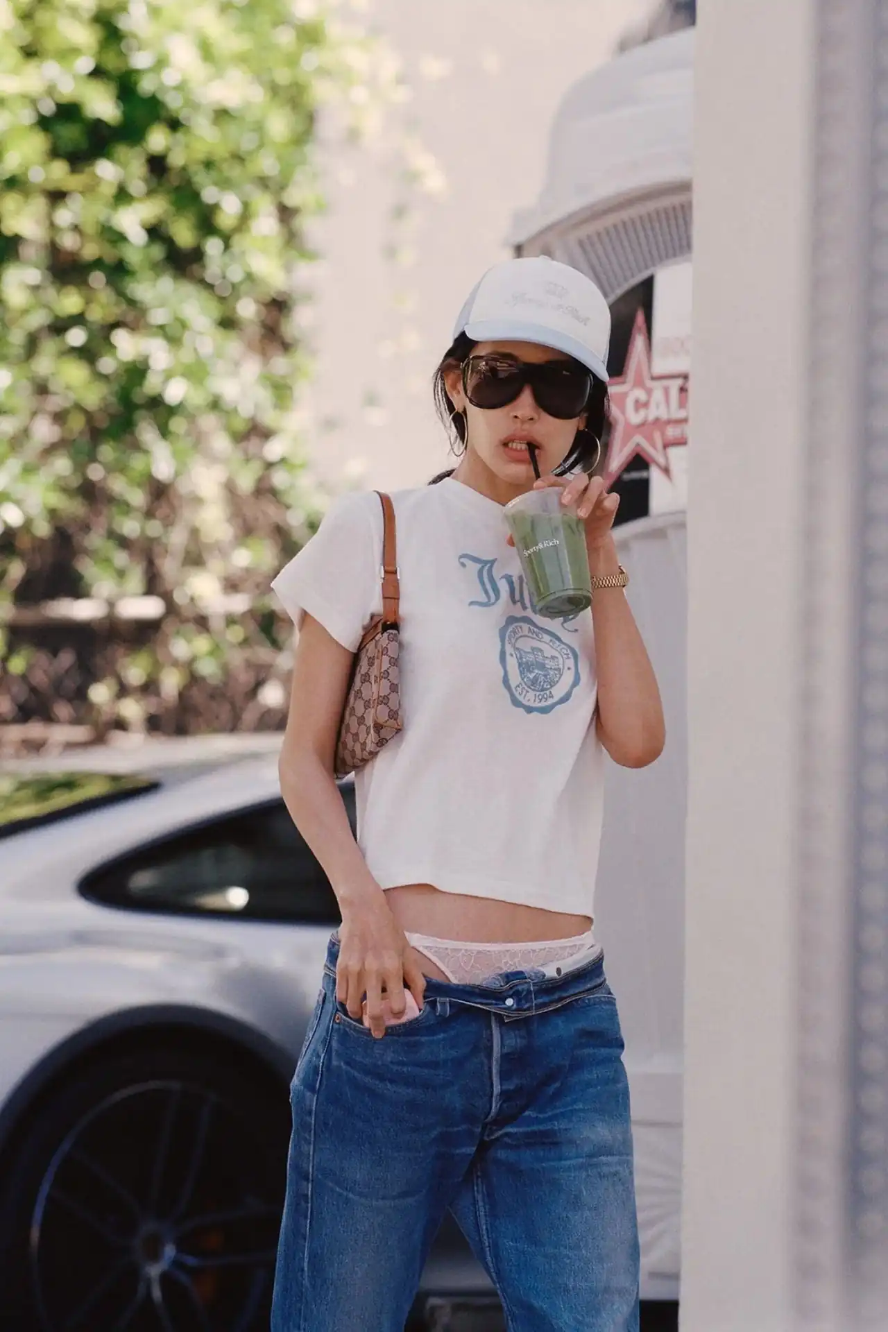 A medium shot shows a person wearing a white t-shirt with blue text that reads 'Sporty & Rich EST. 1994' and blue jeans, holding a green drink and a s...