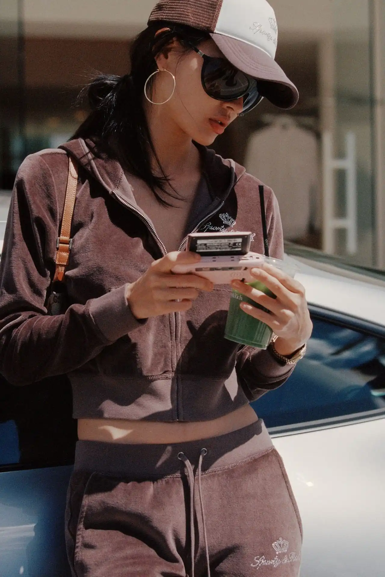 A medium shot captures a person wearing a brown velour tracksuit and a brown and white trucker hat, holding a pink device and a green drink. The track...