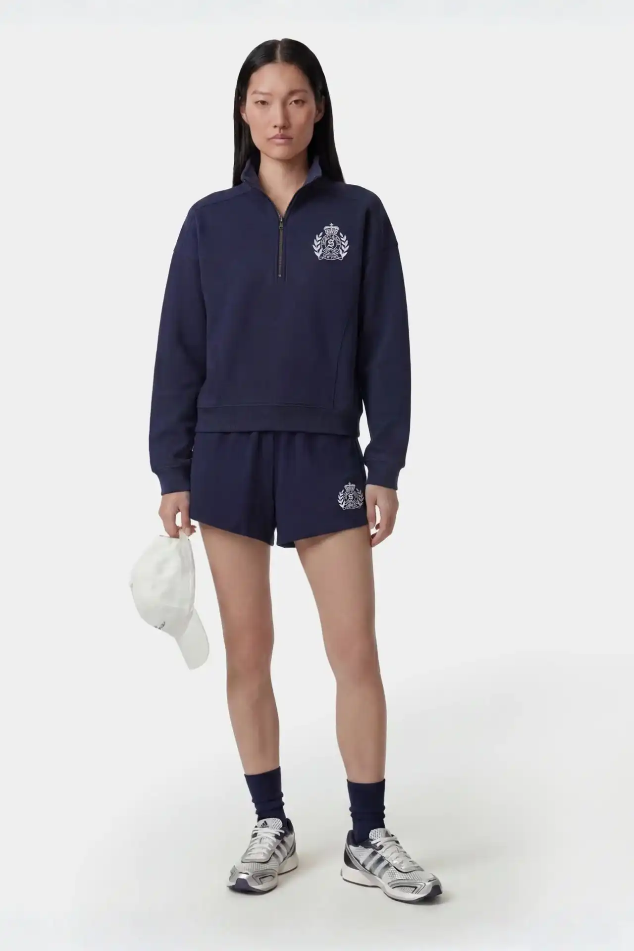 A medium shot shows a woman standing against a white background, wearing a navy blue quarter-zip sweatshirt and matching shorts, both adorned with a w...