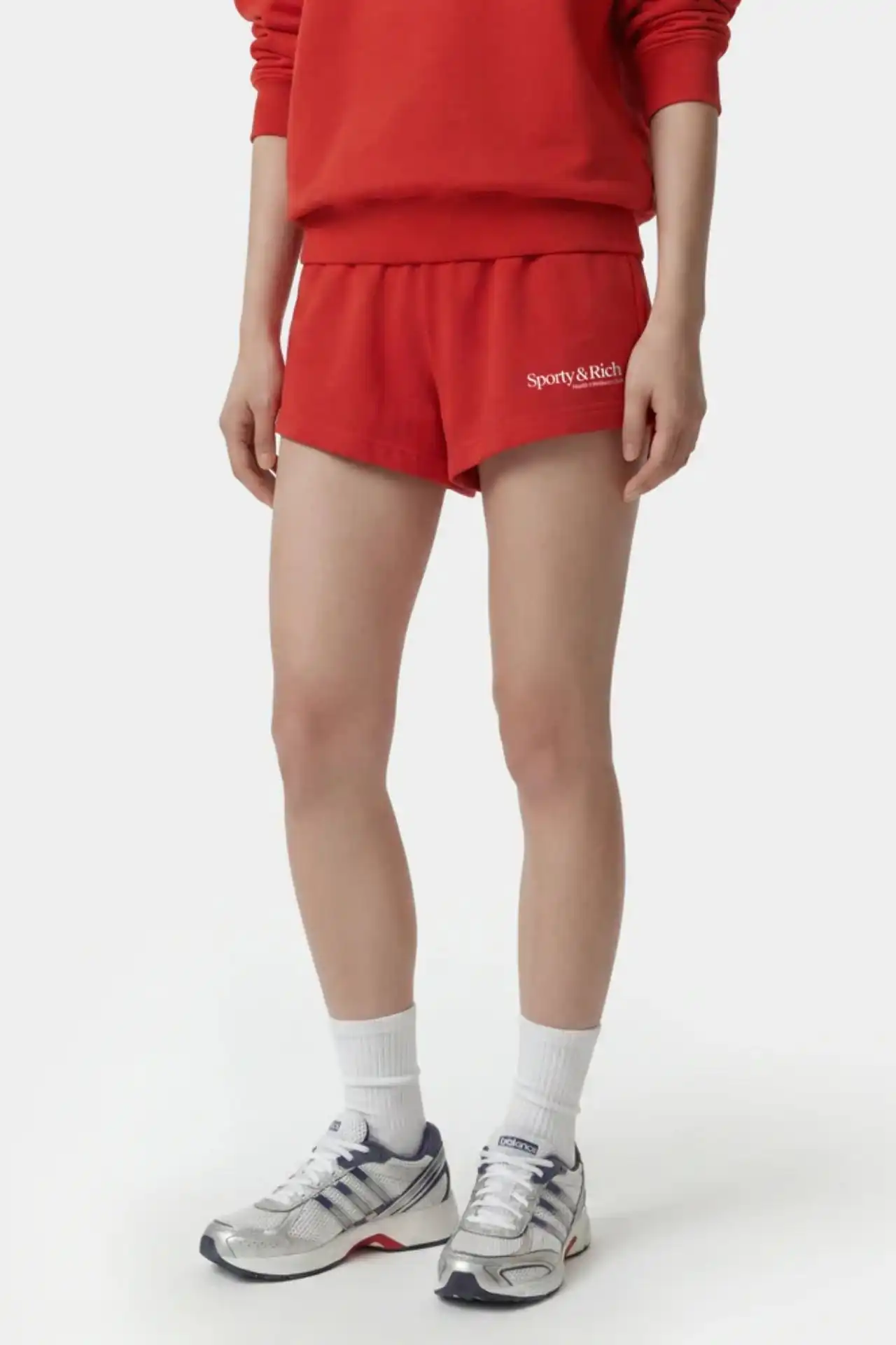 A medium shot shows a person wearing red athletic shorts with the white text 'Sporty & Rich' printed on the left leg, a red sweatshirt, white socks, a...