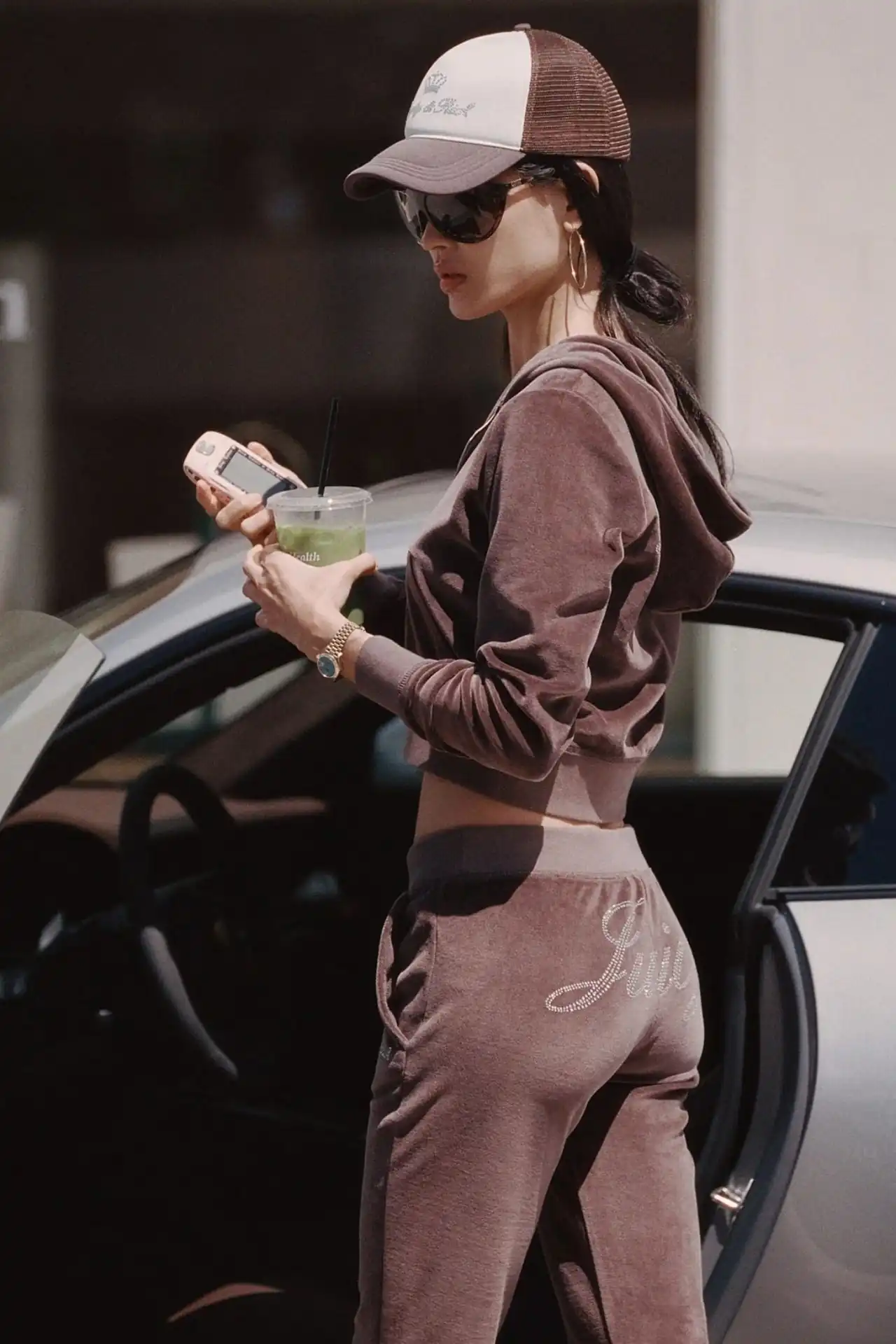 A medium shot shows a person wearing a brown velour tracksuit and a brown and white trucker hat, holding a pink cell phone and a green drink with a st...