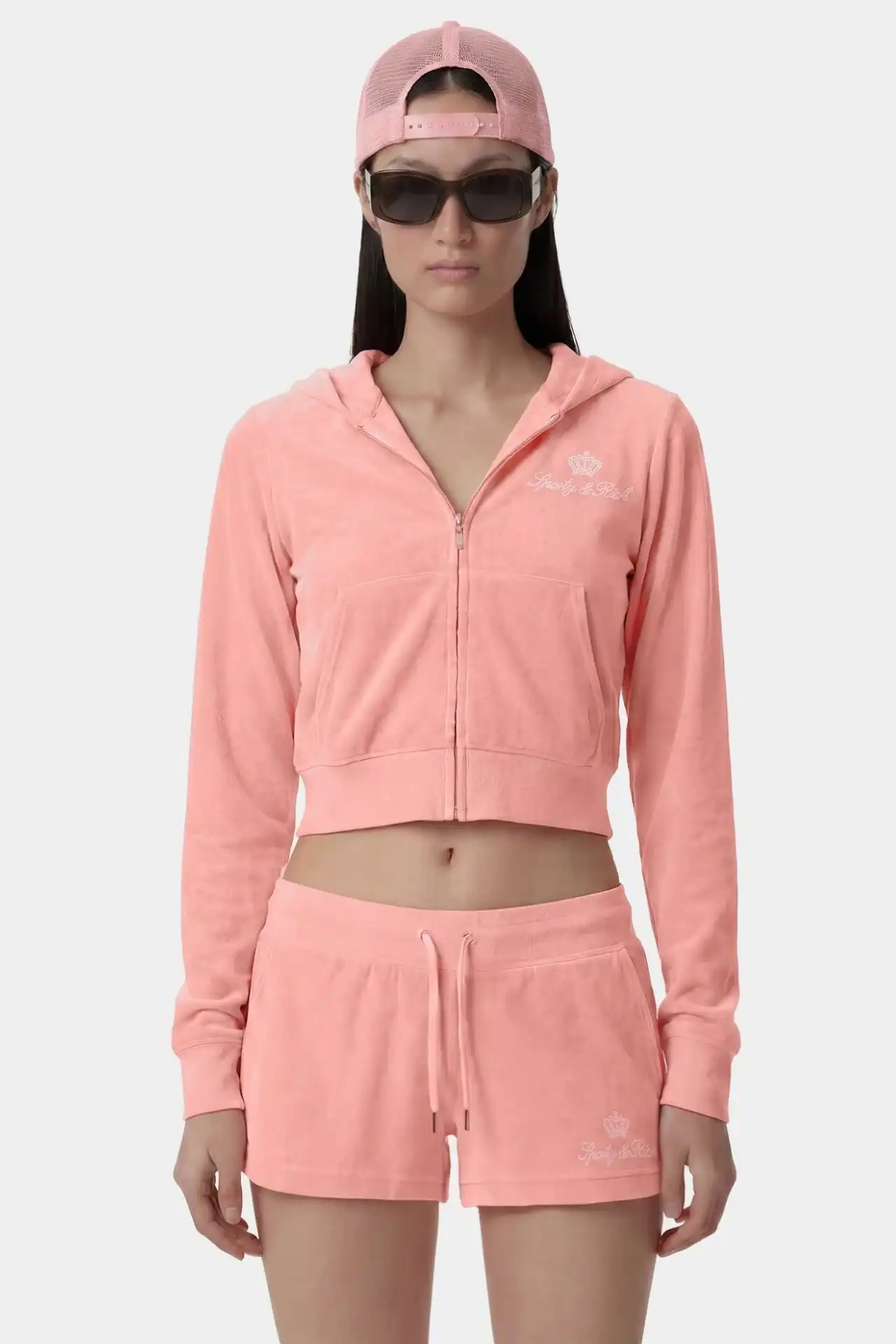 A medium shot shows a person wearing a pink velour tracksuit consisting of a cropped zip-up hoodie and shorts, with a pink mesh trucker hat and dark s...