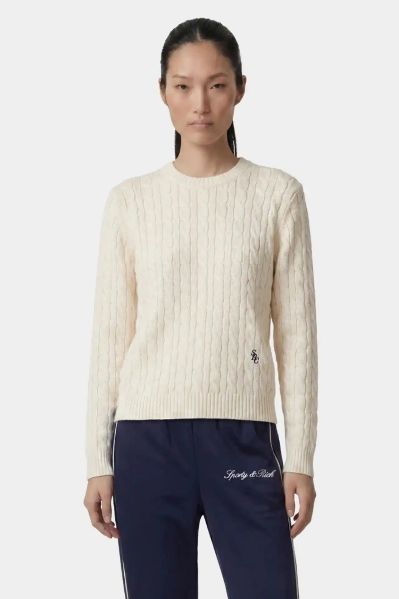 A medium shot shows a woman from the chest up, wearing a cream cable-knit sweater with a small navy blue 'SC' logo on the left chest and navy blue tra...