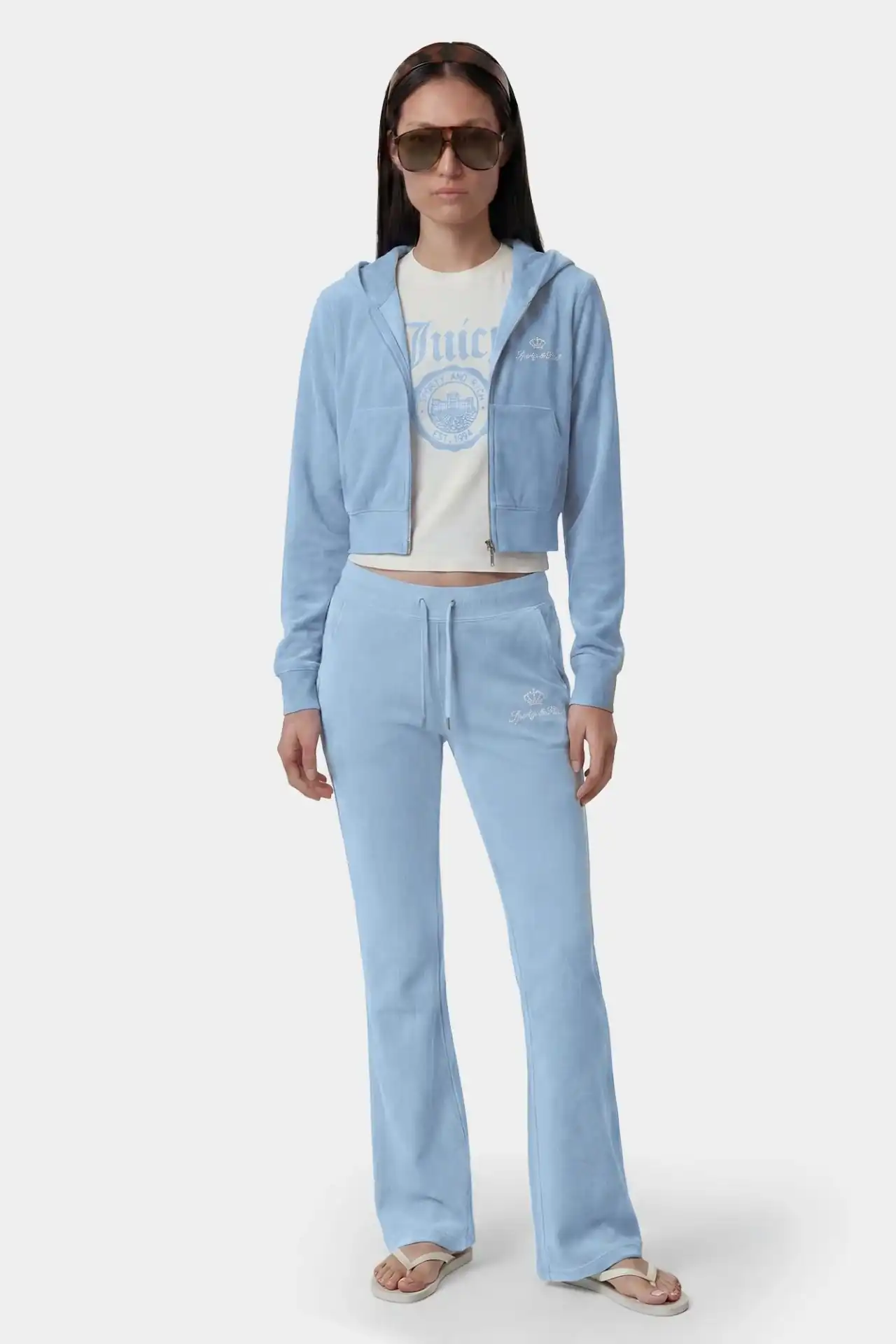 A medium shot shows a woman wearing a light blue velour tracksuit consisting of a cropped zip-up hoodie and flared pants, with a white t-shirt underne...