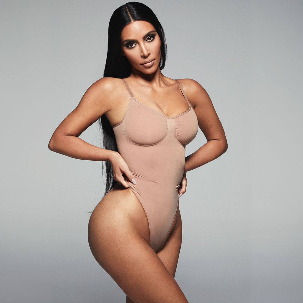 A woman with long dark hair poses confidently in a beige bodysuit against a plain background.