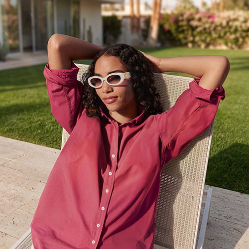 A woman in a red shirt and white sunglasses lounges on a chair outdoors, relaxing with arms up.