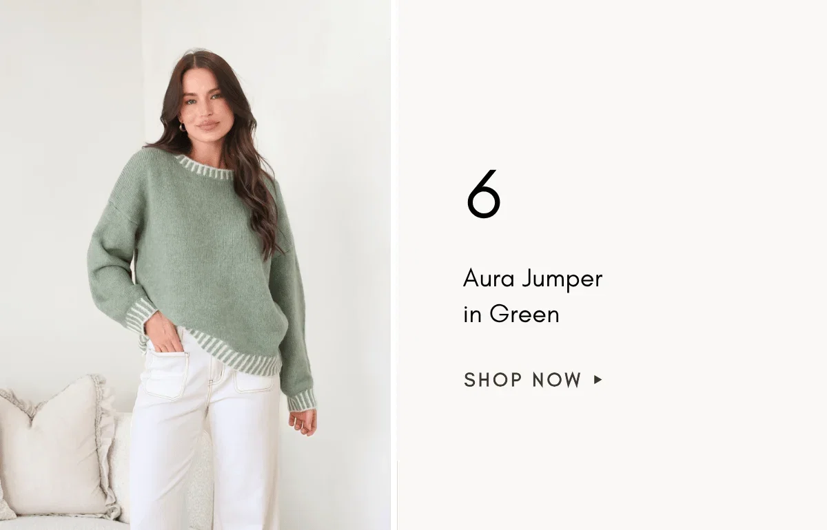 A medium shot shows a woman wearing a green Aura Jumper in Green with white striped trim and white pants, standing against a white background with a c...
