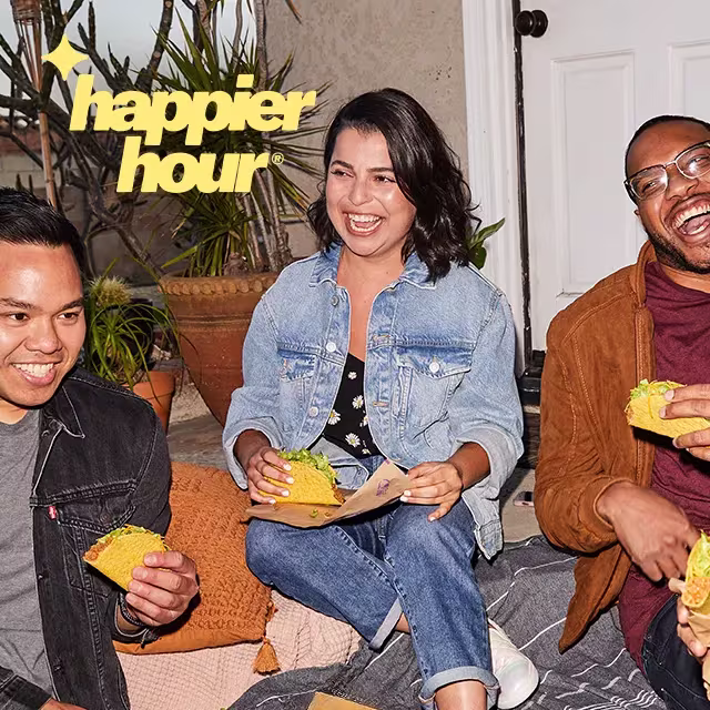 Three friends laugh together outdoors while eating Taco Bell tacos during a "happier hour" gathering.