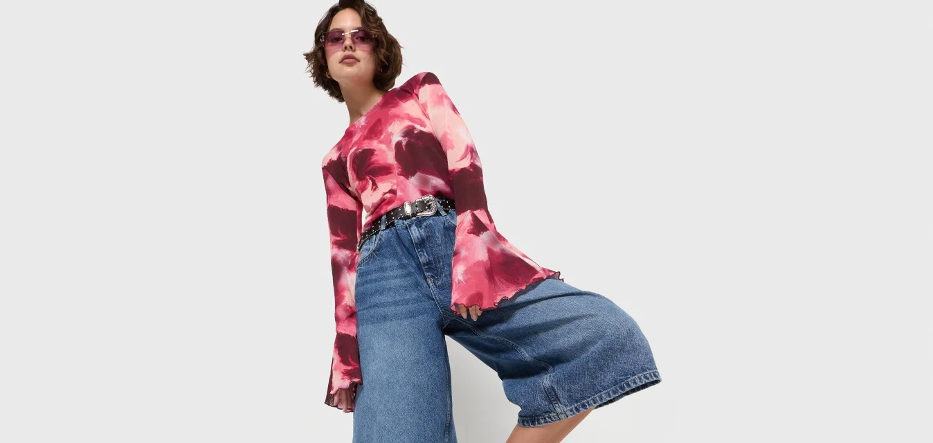 Langekous, rood en roze tie-dye shirt, blauwe denim culottes en zwarte sandalen met gespdetails. Eenvoudige achtergrond benadrukt de outfit.