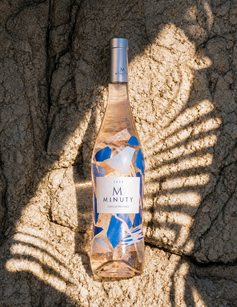 Bottle of 2024 Minuty rosé with blue abstract label lying on sunlit textured rock, palm-leaf shadow