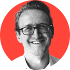 A black and white headshot of Mitchell Labiak set against a red background. He wears glasses, a white shirt and is smiling.