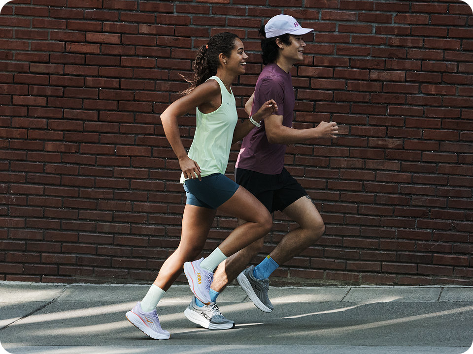 Image of a woman and a man running side by side. There is a red brick wall in the background.
