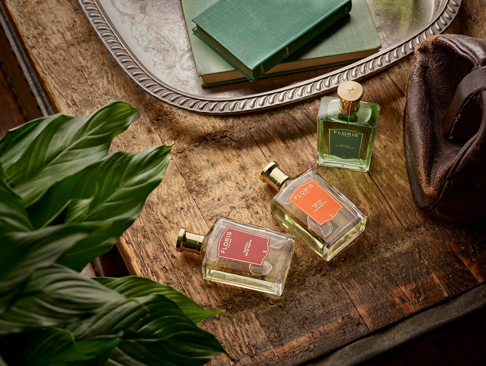 Three bottles of Floris perfume rest on a rustic wooden table beside a green plant, a silver tray with books, and a rustic brown leather bag.