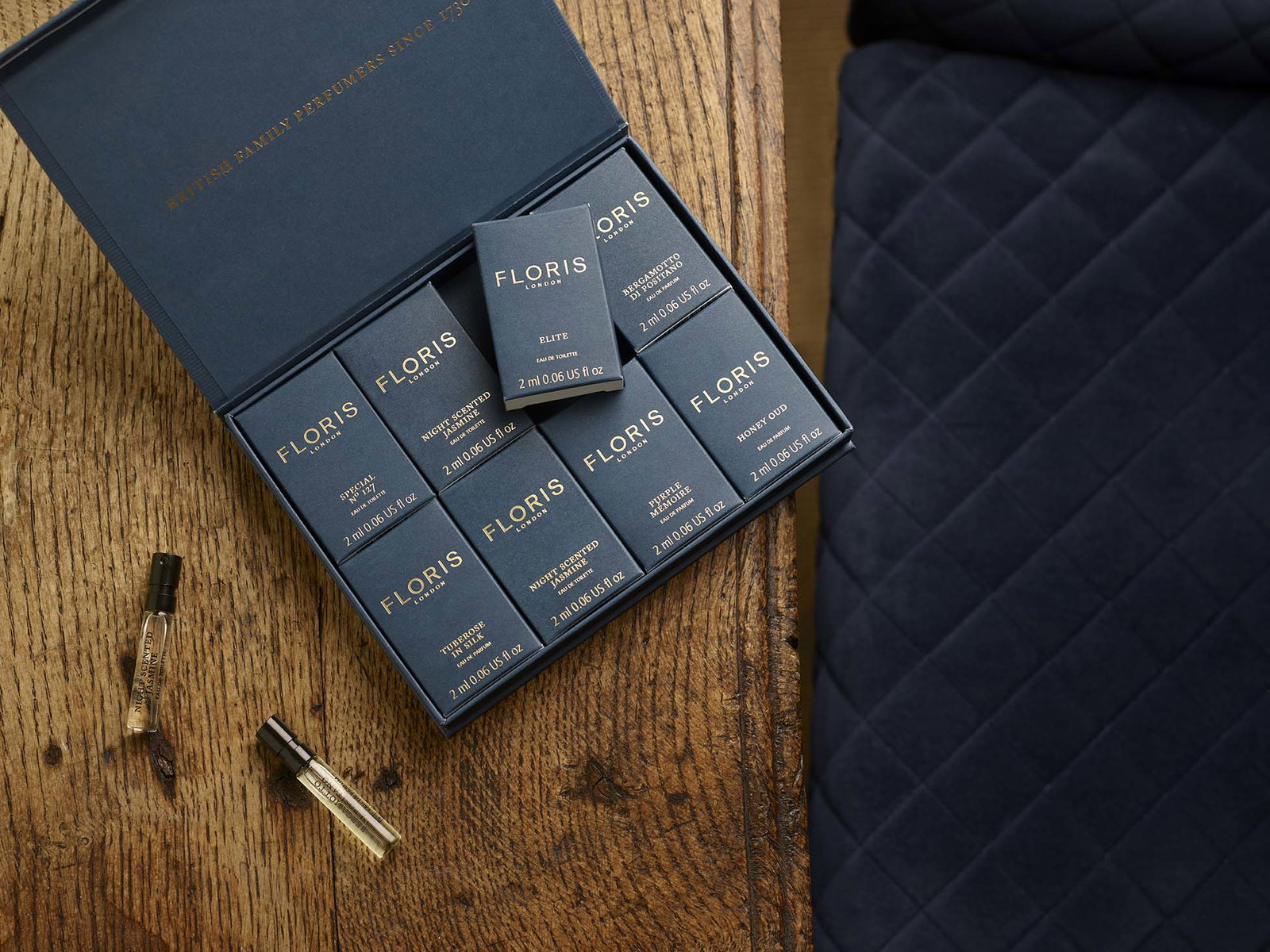 A navy blue Floris perfume sample box sits open on a wooden table next to two small perfume vials, with a quilted navy chair nearby.