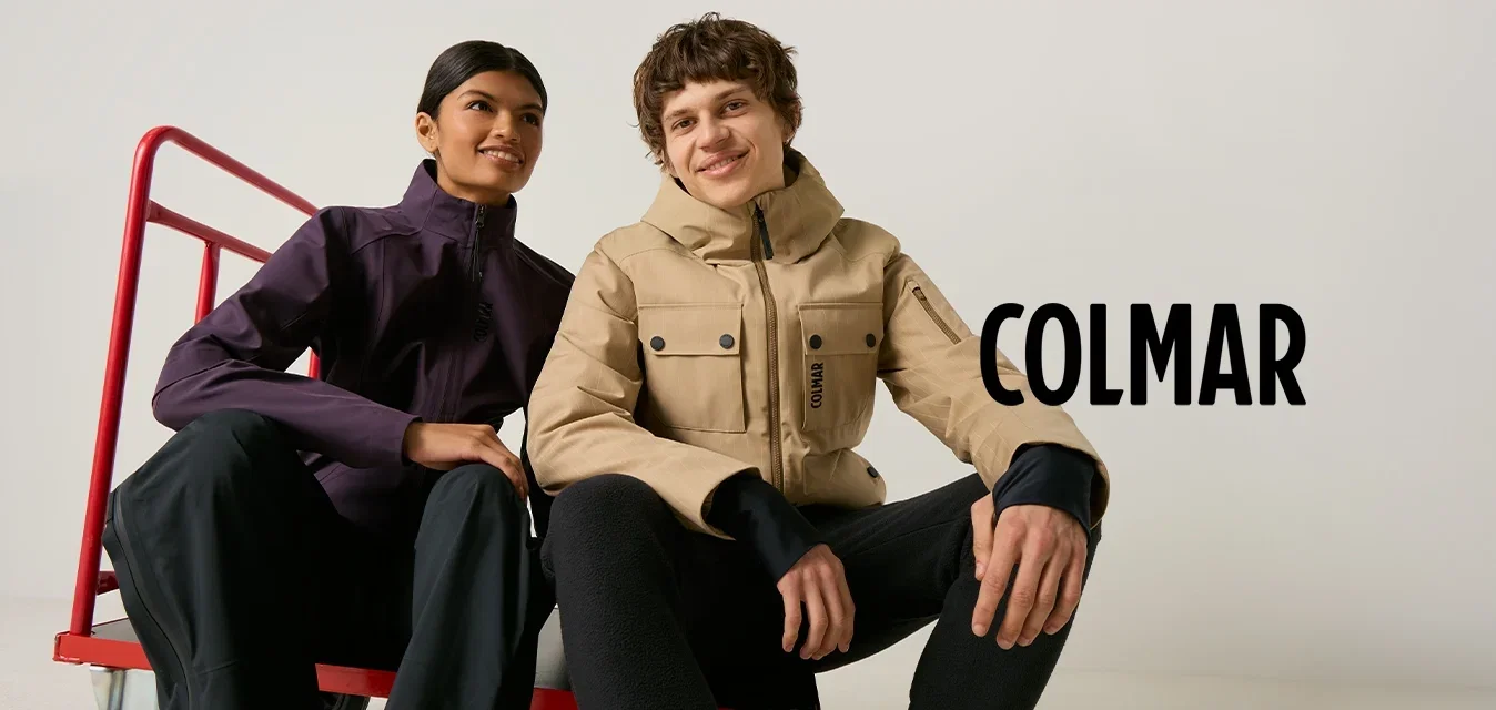 Dos jóvenes adultos están sentados sobre un carrito de mano rojo, vistiendo chaquetas casuales para exteriores y pantalones negros, sonriendo frente a un fondo liso.