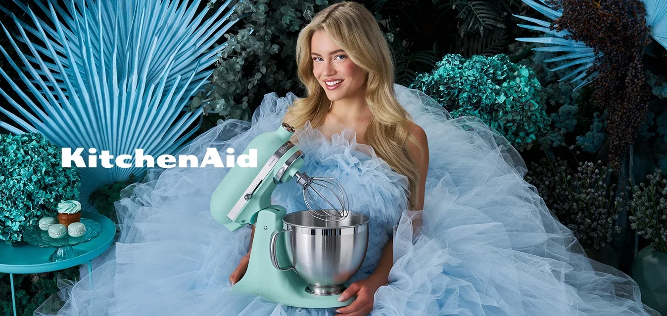 Femme dans une robe ample en tulle bleu clair tenant un batteur sur socle vert menthe, entourée de feuillage bleu-vert et de cupcakes sur une petite table.
