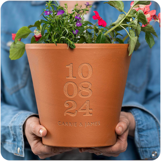 A pair of hands holding an engraved terracotta pot with a small flowering plant, personalised with a special date