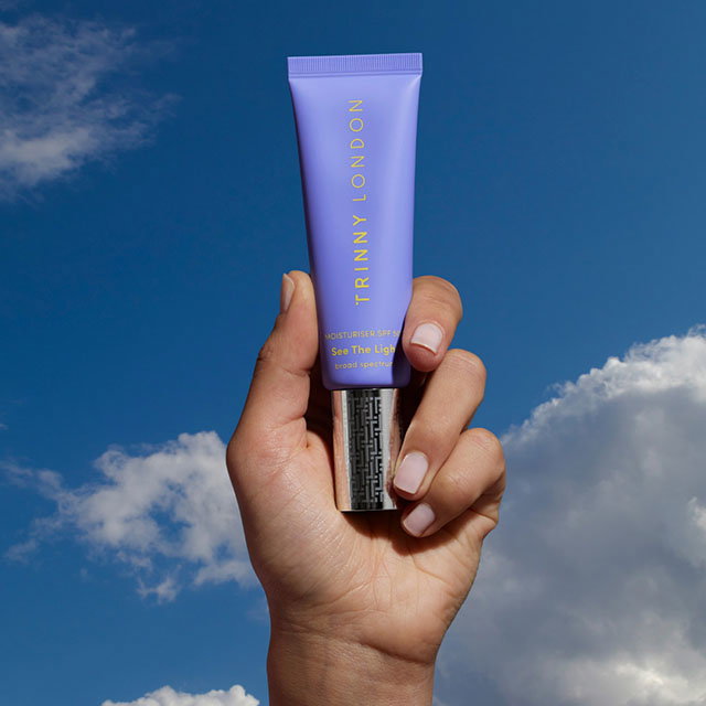 A hand holds a purple tube of Trinny London 'See The Light' skincare product against a blue sky with clouds.
