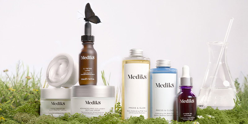 A visually serene and clinical composition showcasing a diverse range of Medik8 skincare products set within a lush, natural environment. The image features various bottles and jars - including the C-Tetra Luxe, Press & Glow daily tonic and Advanced Pro-Collagen Peptide Cream - arranged artfully on a bed of vibrant green moss. A laboratory flask sits to the side, bridging the gap between scientific innovation and nature. Adding a touch of delicate life, a single dark butterfly rests atop the dropper of the C-Tetra Luxe bottle. The clean, white background and soft lighting emphasize the brand's premium, results-driven aesthetic, while the greenery underscores Medik8’s commitment to environmental responsibility and its status as a Certified B Corp™.