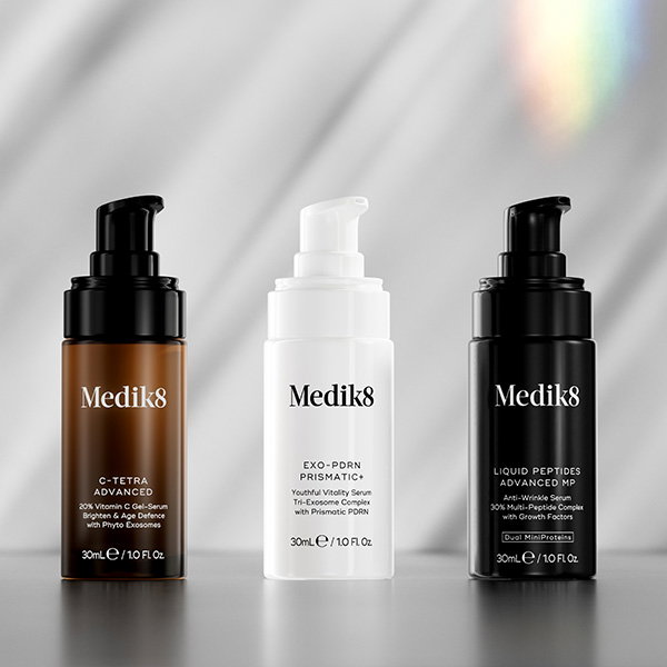 Three skincare pump bottles standing side-by-side against a soft, draped grey fabric background. From left to right: C-Tetra Advanced, Exo-PDRN Prismatic+, and Liquid Peptides Advanced MP.