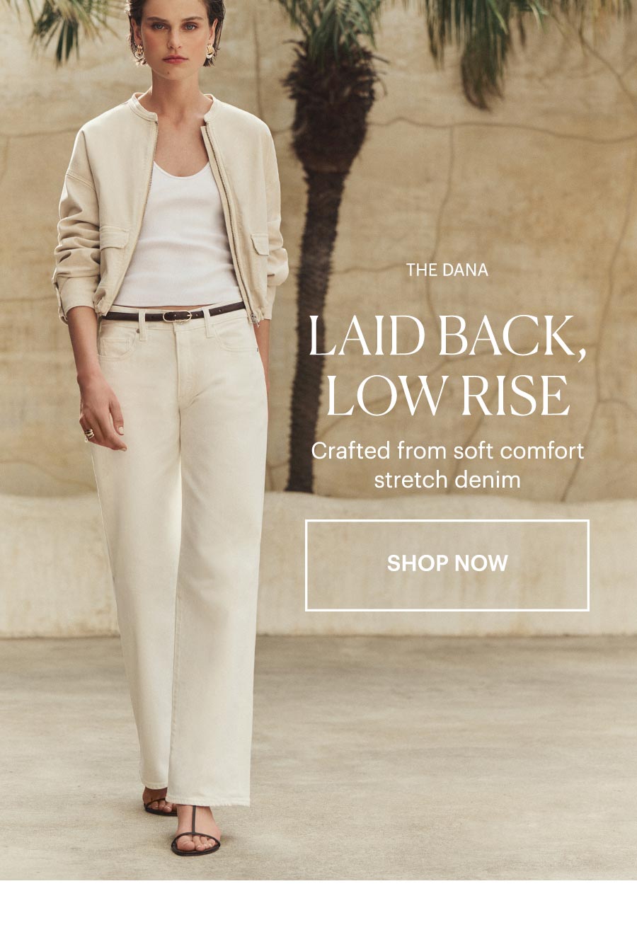 The Dana: Laid Back, Low Rise - Crafted from soft comfort stretch denim. Shop now. Featuring a woman in a cream zip-up jacket, white tank, and pair of low-rise wide-leg ecru jeans, posed in front of a sunlit stone wall and greenery.