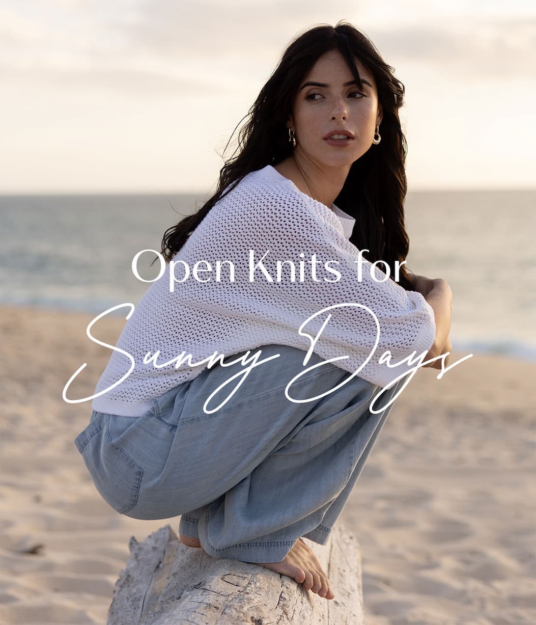 Model wrapped in a white open-knit sweater while sitting on a beach log, with overlay text promoting Open Knits for sunny days.