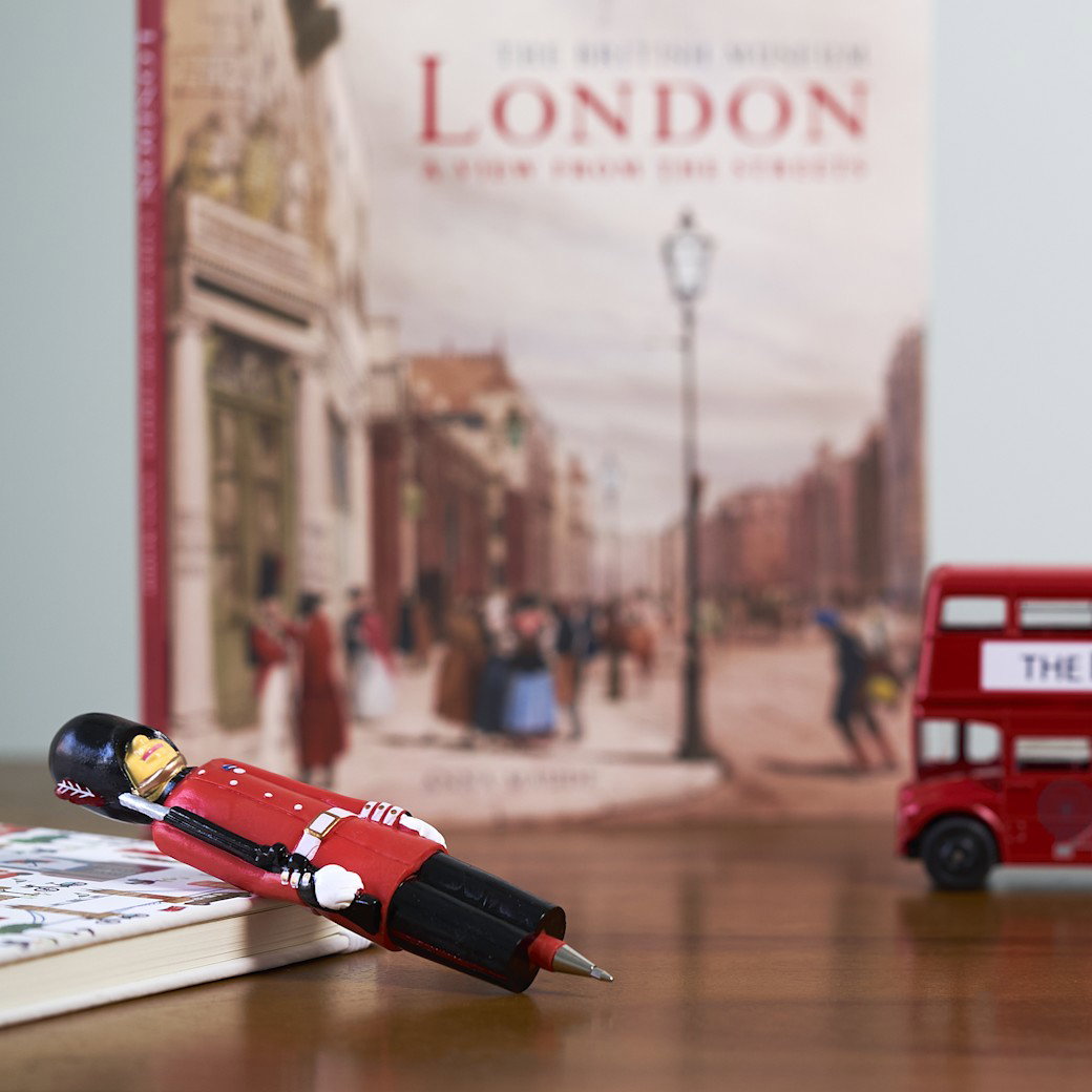 A London guardsman pen, a notebook and a mini-London bus lay on a wooden table in front of a book about London. – Shop London Gifts. 