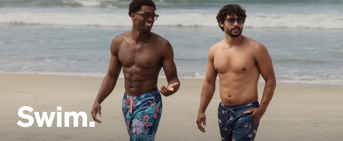 Two models walking on the beach wearing floral SAXX swim shorts with “Swim.” text overlay.