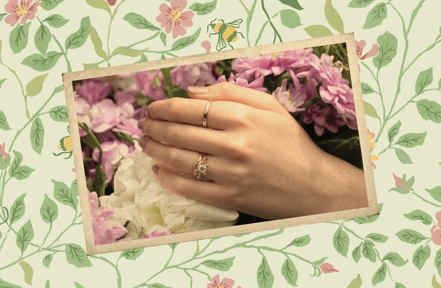 Hand adorned with Alex Monroe floral rings resting among pink hydrangeas and white flowers, set against a colourful backdrop and framed by a floral pattern