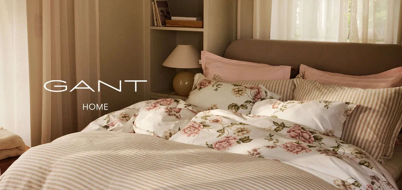 Camera da letto soleggiata con un letto ben rifatto, con biancheria da letto a righe e floreale, tende leggere, una lampada da comodino e uno sgabello piccolo con coperte piegate.