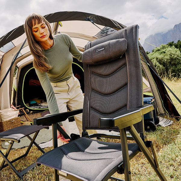 A person sets up a reclining camping chair in front of a large tent on a grassy campsite. Additional folding chairs and camping gear are placed nearby, with trees and hills visible in the background.