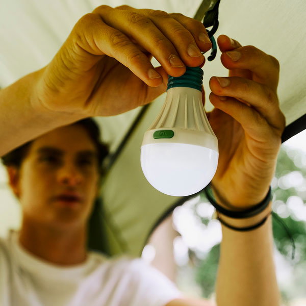 Close-up of hands attaching a portable bulb light to a tent frame using a hook. The hanging light has a green power button and a white diffuser, with the blurred interior of a tent and a person in the background.