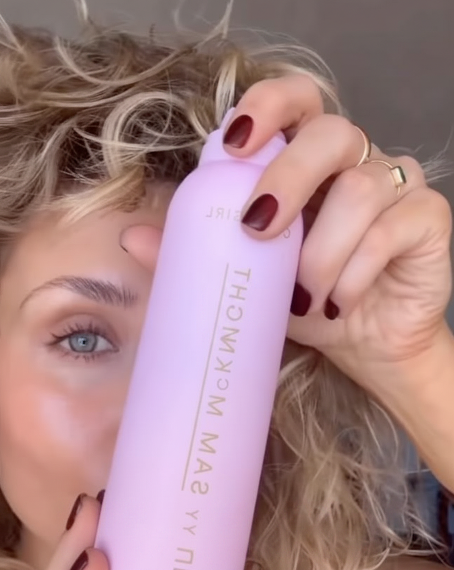 Woman with curly blonde hair holds a pink spray bottle in front of her face.