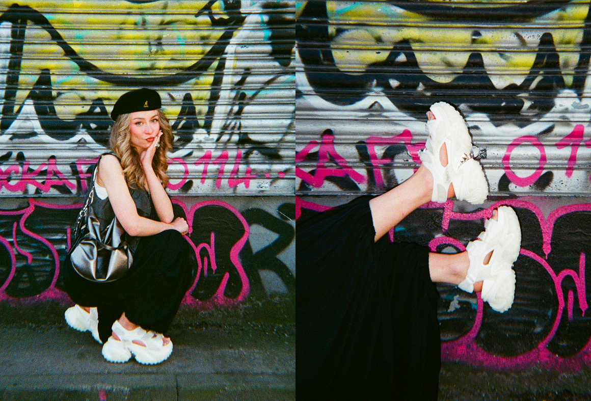 Person sitting against graffiti wall wearing chunky white sneakers and black outfit with shoulder bag