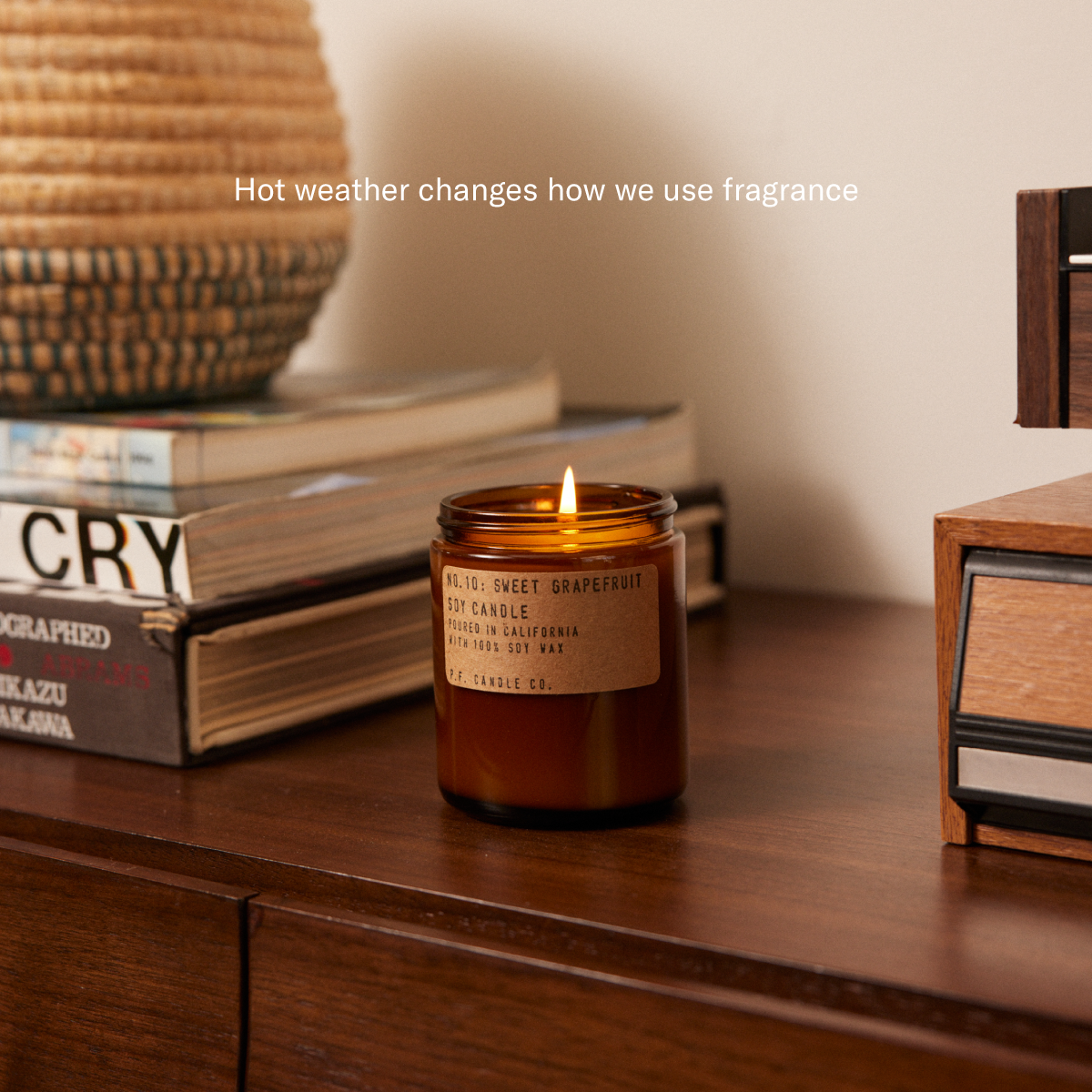 Hot weather changes how we use fragrance [lit candle on wooden surface amongst books, audio equipment, and wicker basket]