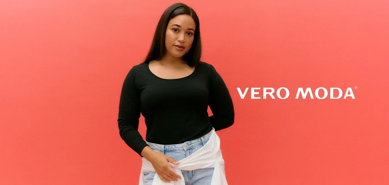 Vrouw met lang donker haar die een zwart shirt met lange mouwen, lichtblauwe jeans en een wit overhemd geknoopt rond haar taille draagt, tegen een koraalkleurige achtergrond.