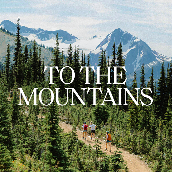 Three people hiking through forest with a snow-covered mountain range in the background. Text overlay reads 'To the mountains.'