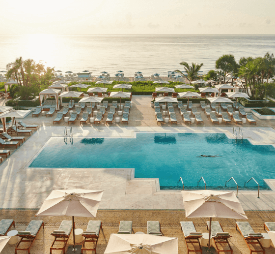 Resort pool with sun loungers and umbrellas facing a sunrise over the ocean.