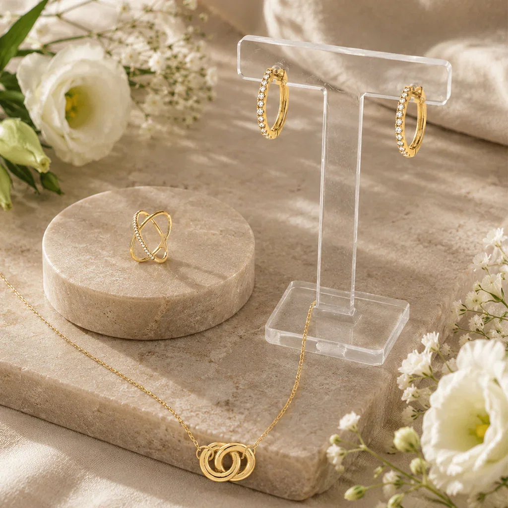 Gold jewelry collection displayed on cream stone platforms with white flowers, featuring diamond hoop earrings, crystal ring holder, and interlocking pendant necklace.