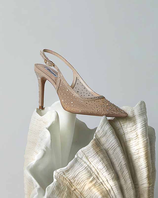 A studded Dune beige high-heeled shoe with an ankle strap is displayed on a textured, shell-like sculpture.