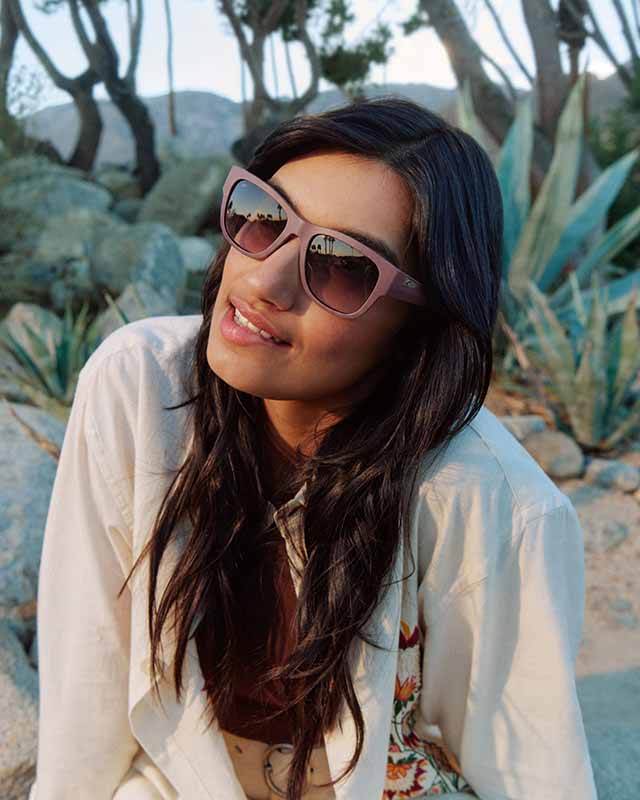 Woman with long hair wearing pink sunglasses and a light jacket, outdoors with desert plants and rocks in the background.