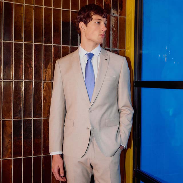A man in a beige suit and blue tie stands near a tiled wall and window, looking to his left.