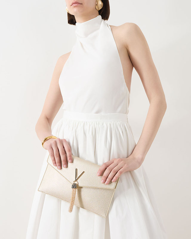 Woman in a white dress holding a Valentino beige clutch with gold logo emblem.
