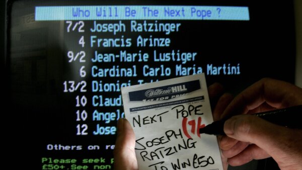 A screen that asks the question of who will be the next pope with names underneath. In front of the screen is a person’s hands holding a puece of paper with their guess. 