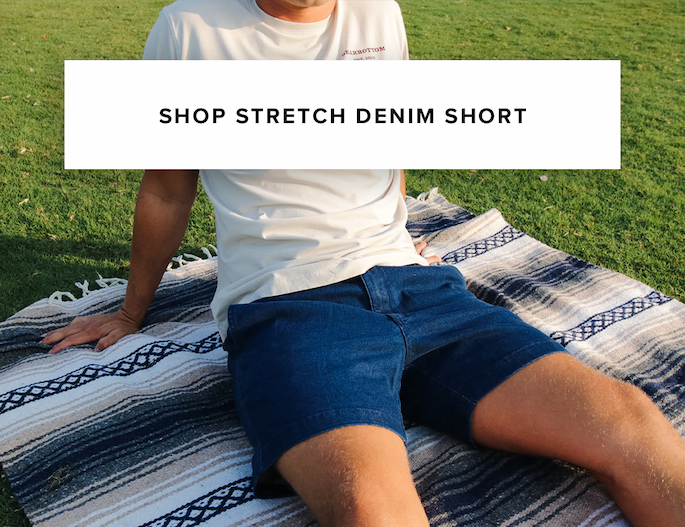 A man sits on a blanket over grass wearing dark blue denim shorts. Text reads: 'Shop stretch denim short.'