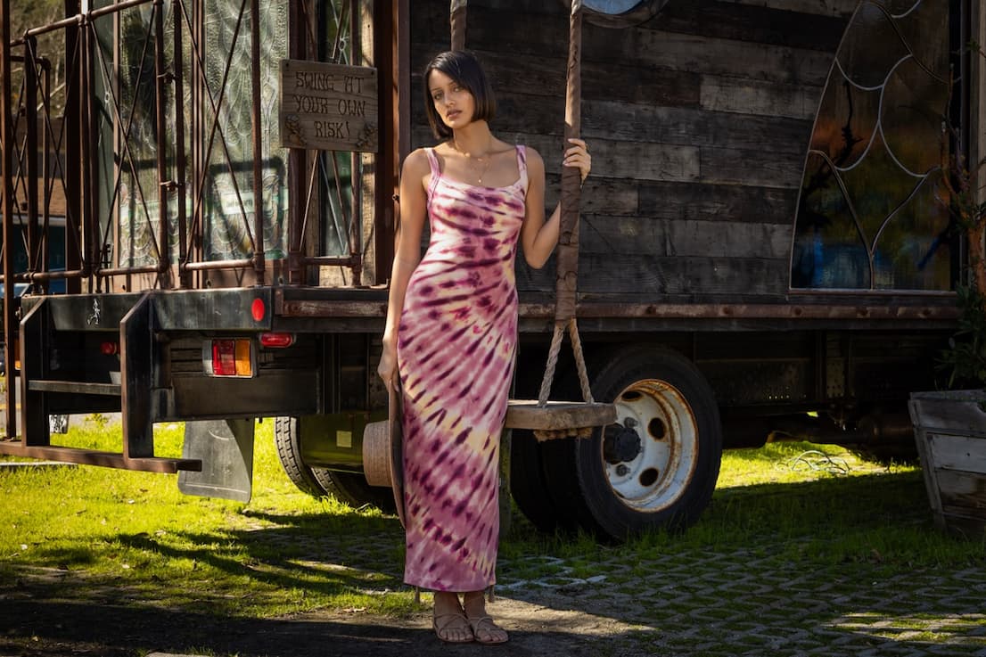Woman wearing a pink tie-dye dress standing outside next to a wood swing