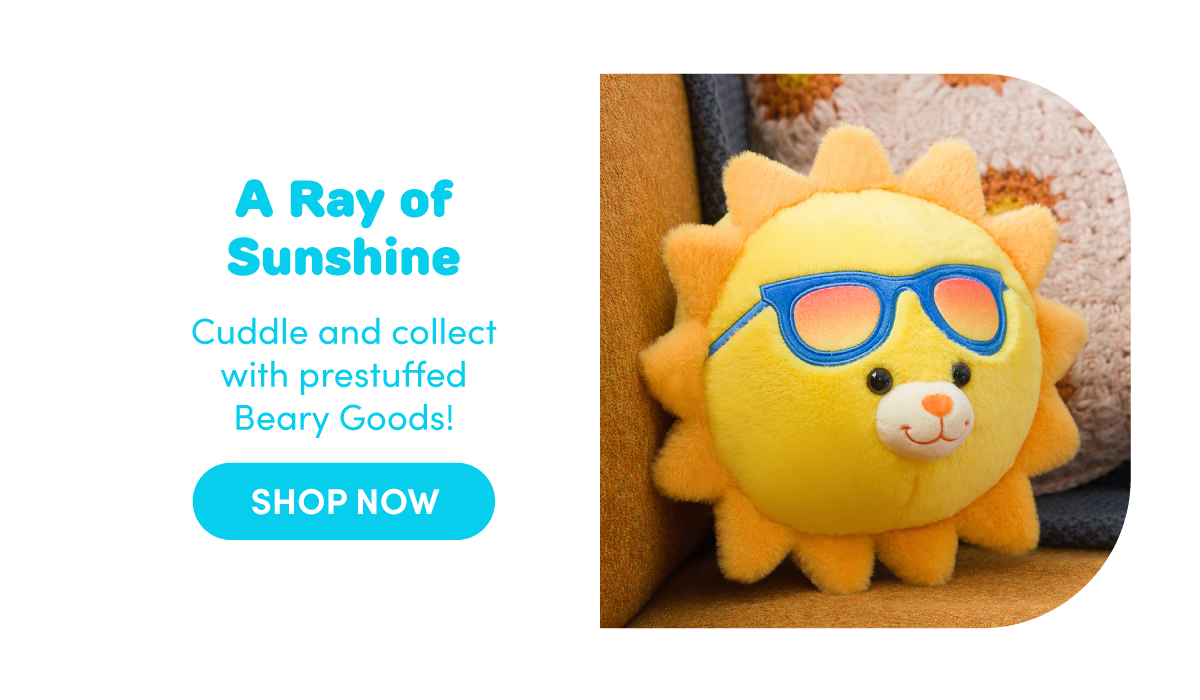Build‑A‑Bear Beary Goods graphic with a split layout on a white background. On the right, a round yellow sun‑themed plush sits against a cozy indoor setting, featuring soft plush rays, a smiling embroidered face, and blue sunglasses with a warm gradient tint. On the left, blue text reads A Ray of Sunshine. Supporting text below reads Cuddle and collect with prestuffed Beary Goods! A rounded blue button beneath the text reads SHOP NOW.