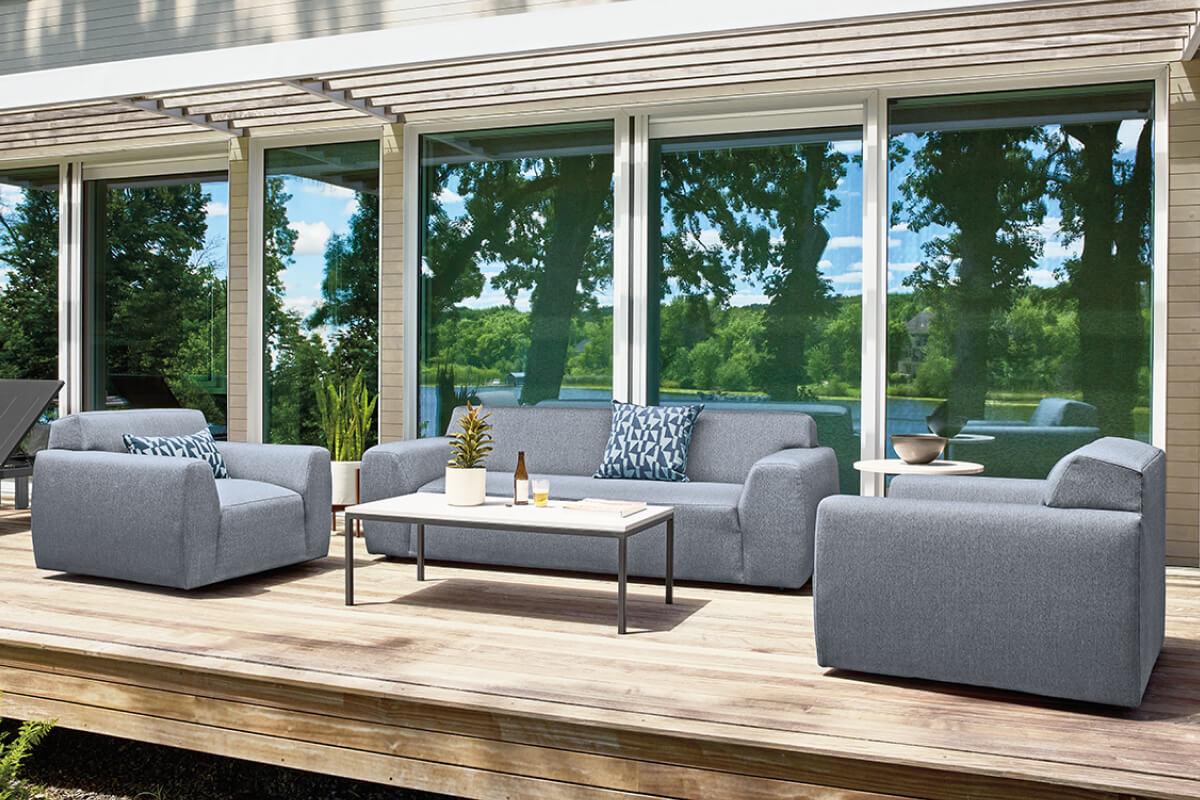 Drift outdoor sofa and chairs in grey upholstery on a wood deck beside floor-to-ceiling lakeview windows.