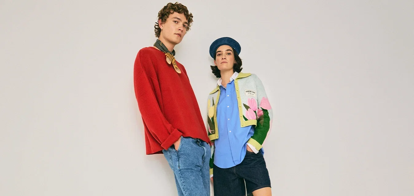 Dos jóvenes adultos vestidos con atuendos casuales y coloridos, de pie frente a un fondo claro y liso con posturas relajadas.